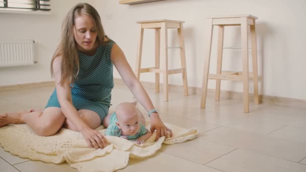 Bella madre sta sorridendo guardando il suo bambino cercando di strisciare su una coperta sul pavimento della cucina. Lento mo — Video Stock