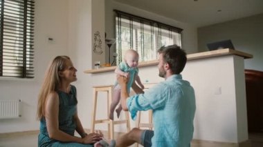 Young mother is sitting on the kitchen with her husband watching him lifting their baby up high. Slow motion
