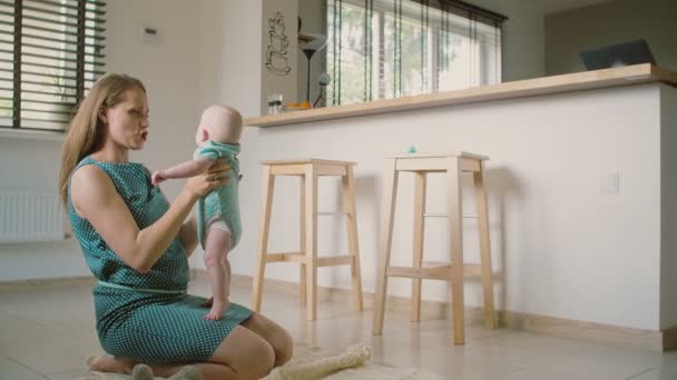 Una hermosa madre joven sostiene a su adorable bebé sonriente ayudándole a ponerse de pie y hablar con él. Movimiento lento — Vídeos de Stock