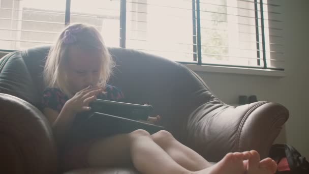 Egy aranyos kislány ül nagy karosszékben az ablak közelében játszik valamit a tabletta. — Stock videók
