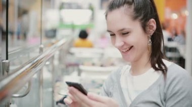 Bir kız bir yan açıdan bakış. Telefonunu kullanarak, gülen ve gülümseyen halka açık bir yerde oturuyor.