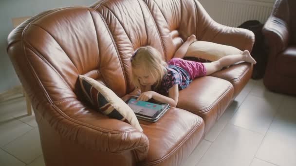Akut sarışın küçük kız tüm pembe giyen kanepede yatıyor ve tablet üzerinde bir oyun oynuyor. Yavaş çekim — Stok video