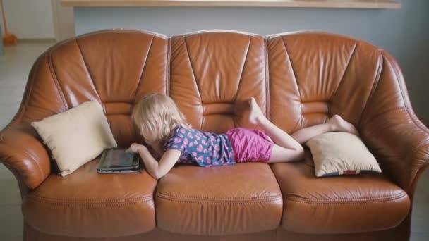 Una vista lateral de una linda niña rubia vestida de rosa está acostada en el sofá y jugando un juego en la tableta. Movimiento lento — Vídeo de stock