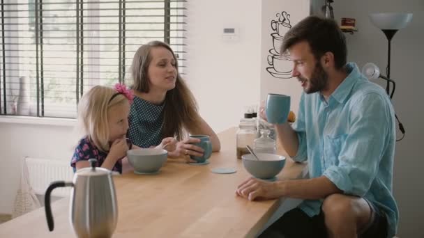 Jovem bela família feliz de três está tomando café da manhã na mesa da cozinha e rindo. Movimento lento, Steadicam tiro — Vídeo de Stock