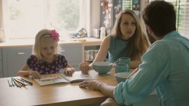 Little girl is showing her colouring book to her parentswhile they are having breakfast at the kitchen table. Slow mo — Stock Video