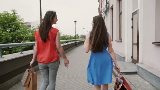 Achteraanzicht van mooie jonge vrouwen wandelen langs de winkels, met boodschappentassen, praten bespreken, slow mo stedicam schot — Stockvideo