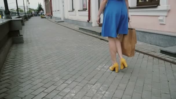 Mooie jonge vrouw in blauwe jurk wandelen in de stad met boodschappentassen, kijkt rond, slow mo stedicam schot — Stockvideo