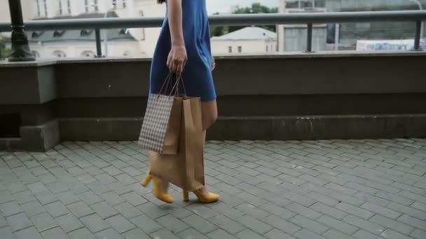 Молодая женщина в синем платье, разгуливающая по городу с сумками для покупок, медленно снимает на камеру — стоковое видео