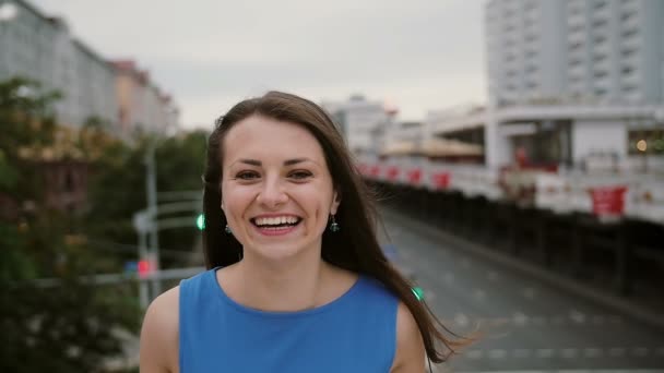 Glimlachend mooi meisje staande op de brug, aanraken van haar haar en kijkt naar de camera. Wind waait haar lang haar. — Stockvideo