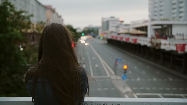 Όμορφο κορίτσι στέκεται στη γέφυρα, κοιτάζει την πόλη, άνεμος φυσάει τα μαλλιά της. πίσω όψη, αργή κίνηση — Αρχείο Βίντεο