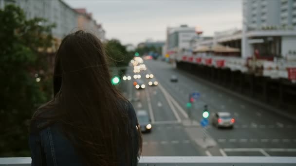 Hermosa chica de pie en el puente, mira a la ciudad, luego se vuelve a la cámara y sonriendo. El viento sopla su cabello. cámara lenta — Vídeos de Stock