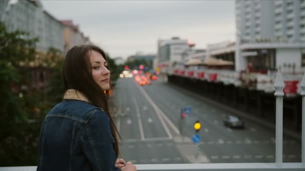 Hermosa chica de pie en el puente, mira a la ciudad, luego se vuelve a la cámara y sonriendo. El viento sopla su cabello. lento mo — Vídeos de Stock