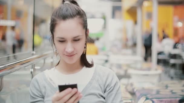 Uma jovem está sentada em um lugar público, usando seu telefone e sorrindo . — Vídeo de Stock