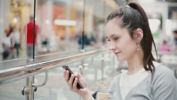 Una vista de una chica desde un ángulo lateral. Ella está sentada en un lugar público, usando su teléfono, sonriendo . — Vídeos de Stock