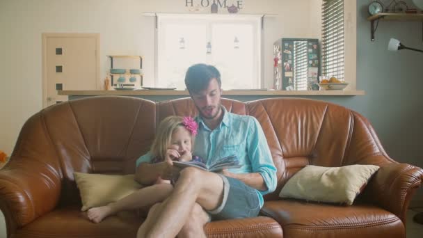 若いハンサムな父親は、彼の貴重な娘に童話を読んでソファーに座っていた。遅い mo ステディカム ショット — ストック動画