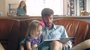 Mutlu bir aile. genç Baba ve küçük sarışın kızı deri kanepe yemek salonunda bir hikaye okuyor. 4k