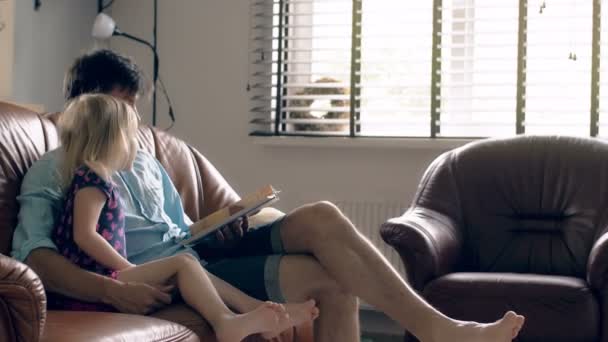 Un joven padre y su pequeña y feliz hija rubia están leyendo una historia sobre un sofá de cuero en el comedor. 4K — Vídeos de Stock