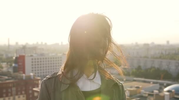 Jeune femme brune debout sur le toit au coucher du soleil tourne la tête et ses cheveux soufflent par le vent. ralenti — Video