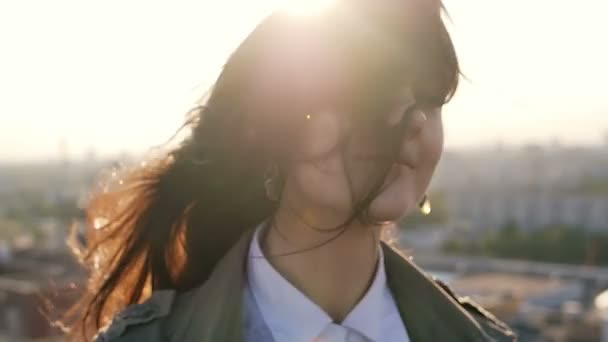 Primer plano. joven mujer de pie en el techo al atardecer gira la cabeza y su pelo está soplando por el viento. lento mo — Vídeo de stock