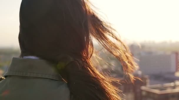 Pohled zpět. Vítr fouká dlouhé tmavé vlasy krásné mladé ženy. dívku stojící na střeše při západu slunce. Zpomalit na mo — Stock video