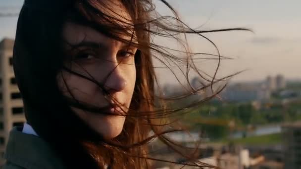 Wind blows long dark hair. girl standing on the roof smiling, looking at the camera. close up. Slow motion — Stock video