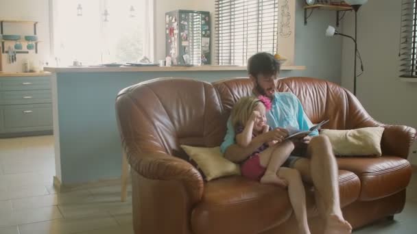En ung stilig far sitter i soffan och läser en bok till sin dyrbara lilla dotter. Långsam Mo, Steadicam shot — Stockvideo