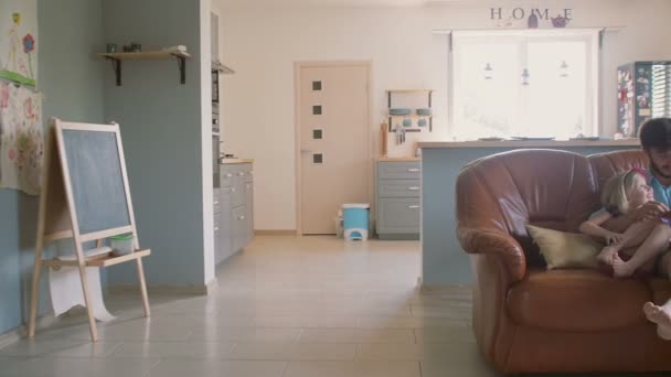 Un joven padre guapo está sentado en el sofá leyendo un libro de cuentos a su preciosa hija. Lento mo, Steadicam tiro — Vídeos de Stock