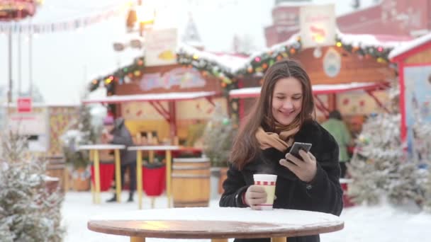 Schöne junge Frau mit Smartphone und heißem Tee während des Weihnachtsmarktes — Stockvideo