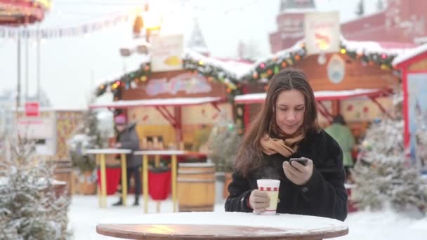 Krásná mladá žena, která používá smartphone a pije horký čaj během vánočního veletrhu — Stock video