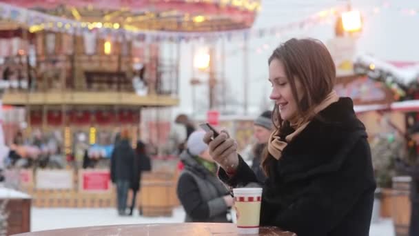 Hermosa mujer joven usando un teléfono inteligente y beber té caliente durante la Feria de Navidad frente al carrusel — Vídeos de Stock