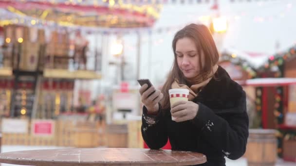 Hermosa mujer joven usando un teléfono inteligente y beber té caliente durante la Feria de Navidad frente al carrusel — Vídeos de Stock