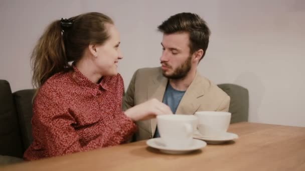 A couple in love sitting on a sofa at the table with empty coffee cups on it. They are talking and smiling, kissing. — Stock Video