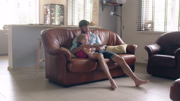 Šťastná rodina. mladý otec a jeho malá blonďatá dcera čtení příběh na kožené pohovce v jídelně. 4k — Stock video