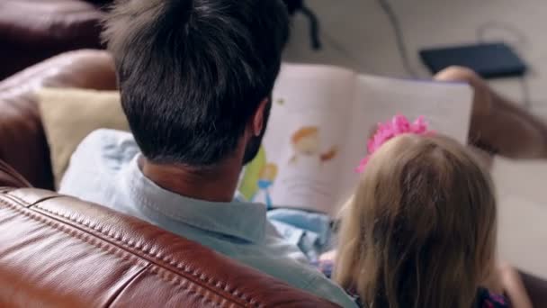 Vista superior. O jovem pai e a sua filha loura estão a ler uma história no sofá de cabedal na sala de jantar. 4K — Vídeo de Stock