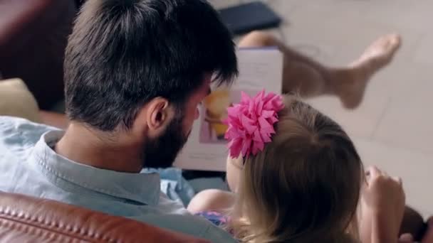 Vue de dessus. le jeune père et sa petite fille blonde lisent une histoire sur un canapé en cuir dans la salle à manger. 4K — Video