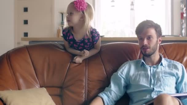 Junge Väter sitzen auf dem Sofa und unterhalten sich mit jemandem, während seine Tochter im Hintergrund lächelt. 4k — Stockvideo