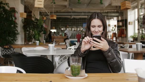 Elegante mujer joven con té de hierbas utiliza un teléfono inteligente sentado a la mesa en la cafetería moderna, sonriendo. 4K — Vídeos de Stock