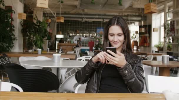 Elegante giovane donna utilizza uno smartphone navigare in internet seduto a un tavolo in un caffè moderno, sorridente. 4K — Video Stock