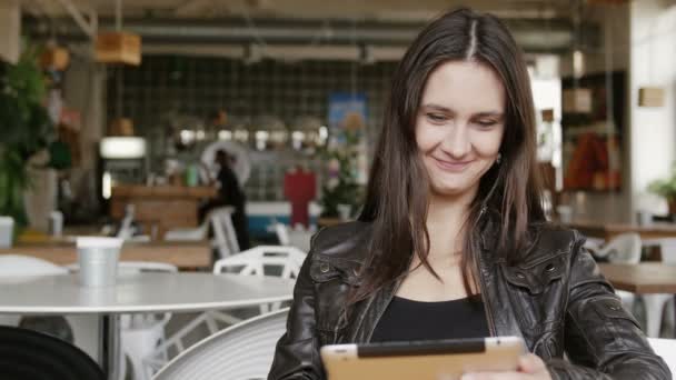 Die elegante junge Frau nutzt ein Tablet und schickt SMS an einen Tisch in einem modernen Café, fragt sich und lächelt. 4k — Stockvideo