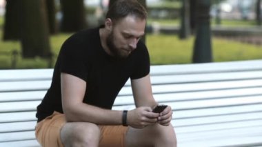 parkta bir bankta oturmuş ve bir smartphone kullanarak yakın çekim gülümseyen çekici genç adam sms gönderir