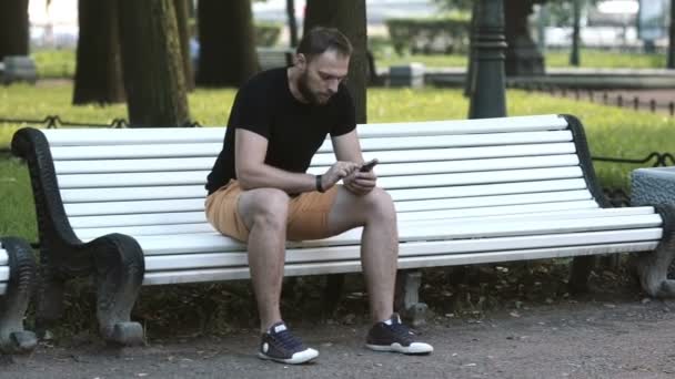 Uśmiechający się atrakcyjny młody człowiek siedzi na ławce w parku i przy użyciu smartfonu — Wideo stockowe