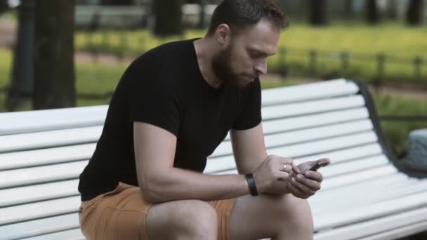 Γκρο πλαν ελκυστικός νεαρός άνδρας κάθεται σε ένα παγκάκι στο πάρκο και χρησιμοποιώντας ένα smartphone — Αρχείο Βίντεο