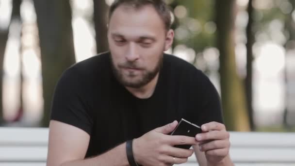 Porträt eines attraktiven jungen Mannes, der auf einer Bank im Park sitzt und ein Smartphone benutzt — Stockvideo