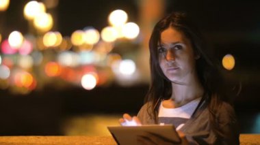 Genç ve güzel kadın tablet kullanır. Görünüyor kamera, gülümseme, göz kırpma, saçlarını dokunur. Gece şehir, bulanık ışıklar, yavaş mo