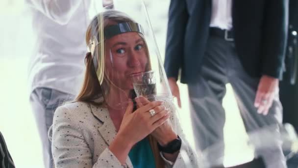 Zabawny portret młodej, szczęśliwej, 30-letniej blondynki biznesowej noszącej tarczę do twarzy, próbującej pić patrząc przed kamerą w biurze. — Wideo stockowe