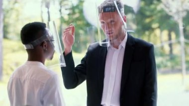 Modern ofis penceresinde konuşan, COVID-19 güvenlik kalkanı takan iki mutlu erkek çoklu etnik iş ortağı.