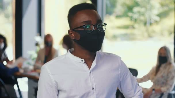 職場の安全。オフィススローモーションで顔マスクを身に着けている眼鏡で若い深刻なアフリカのビジネスマンの肖像. — ストック動画