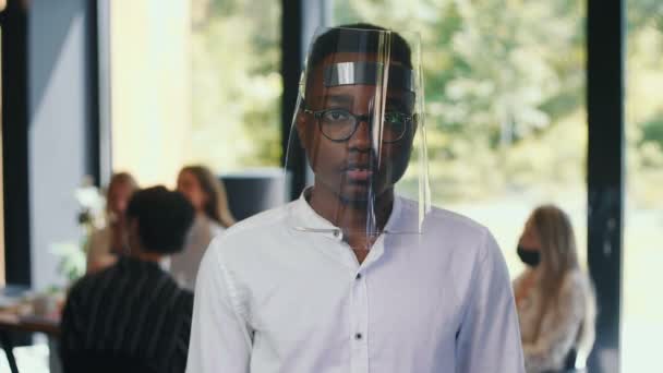 若い深刻な20代のアフリカのオフィスマネージャーの男性の肖像は、光の職場で遅い動きでプラスチック製の顔シールドノードを着用 — ストック動画