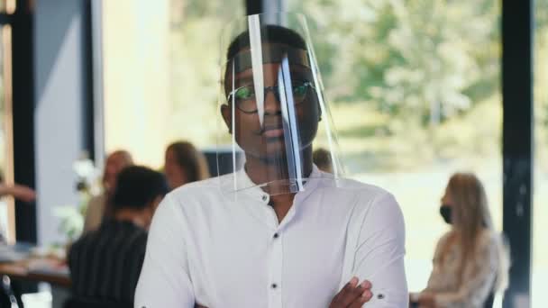 Seguridad en el lugar de trabajo. Retrato del joven empresario africano de 20 años en escudo facial de plástico sonriendo a cámara lenta de la oficina de luz. — Vídeos de Stock