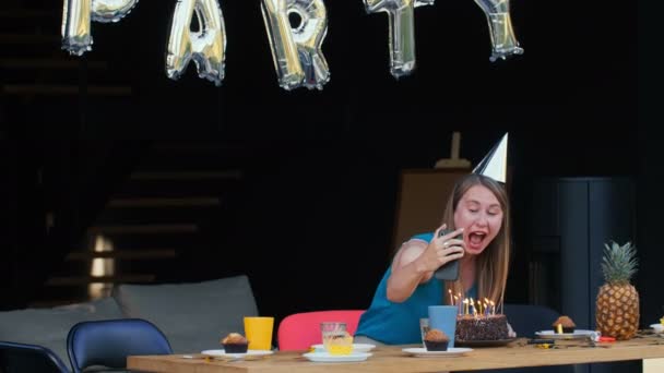 Anniversaire de l'isolement. Heureuse jeune femme caucasienne parle à des amis, souffle sur le gâteau sur smartphone appel vidéo à la maison. — Video